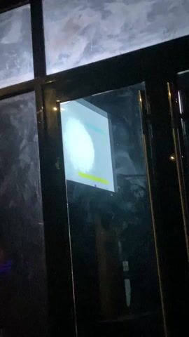 The video is a dark, low-quality recording of what appears to be a reflection in a glass door or window. Inside, a figure resembling a person wearing a light-colored shirt or jacket can be seen. The illumination is poor, but the clothing seems to have a yellow or light-colored stripe, or design, on it. Behind the figure is an indiscernible background, mostly black. Some movement is indicated within the frame, but the quality makes identifying the specific actions of individuals or objects difficult. Towards the very end, the camera moves, ending focused on plain black.