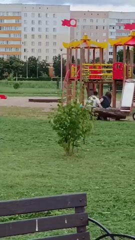The video shows a park scene with a playground in the background. A small tree is visible in the foreground. Further back, there are several umbrellas providing shade over benches, a trash can, and some small white structures that appear to be gazebos or shelters. In the distance, apartment buildings are visible. The sky is cloudy. In the initial frames, two individuals are sitting on a log near the playground. The camera pans across the park, focusing on the umbrellas and the green grass. No specific actions or interactions are clearly discernible beyond the initial presence of the two individuals.
