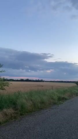 The video captures a scenic view of a rural landscape during what appears to be dusk or dawn. The sky is a gradient of soft blues and grays, with scattered clouds tinged with the warm hues of the setting or rising sun. In the foreground, a paved road or path runs diagonally across the frame, with a rough texture suggesting it might be asphalt or gravel. To the left of the road, tall, dry grass and some greener vegetation form a natural border. Beyond this, a field stretches out, possibly a harvested crop or dry grassland, with a few trees and distant structures visible on the horizon. The overall atmosphere is serene and peaceful, showcasing the beauty of the natural environment. There are no people or animals visible in the footage.