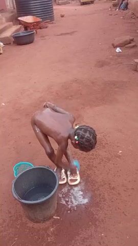 A young person, likely a child, is taking a bath outdoors in a reddish-brown dirt yard. They are standing with their back to the camera, bent over, and using a blue sponge to wash themselves. They are wearing white sandals and their hair is braided with colorful beads. A black bucket full of water sits to their left, and a small blue basket is beside it. In the background, there's a wheelbarrow, a large black tub, various objects scattered on the ground, and parts of buildings.