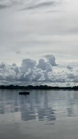 A small boat with two people is visible on a calm body of water. The boat is in the middle ground of the image, and the people are silhouetted against the sky. The water reflects the sky and clouds. The background consists of a tree line and a cloudy sky with dramatic cloud formations. The overall mood of the video is serene and peaceful.