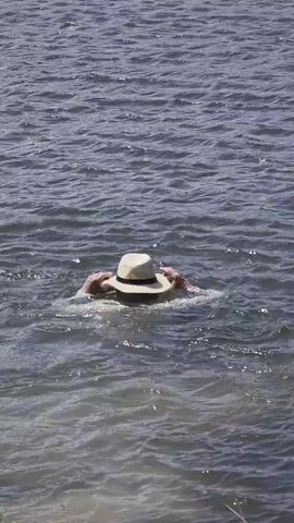 The video shows a person floating face down in a body of water. The person is wearing a hat, a light-colored shirt, and shoes, and their legs are visible near the surface of the water. The water is choppy with small waves, and the shore is visible in the foreground. The video sounds include voices speaking in Russian.