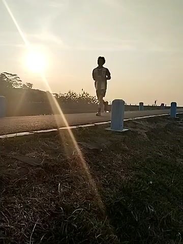 The video features an outdoor scene bathed in the warm light of either sunrise or sunset, with the sun creating a bright glare in the upper left of the frame. A lone figure is visible in the middle distance, silhouetted against the bright sky, jogging or running on a paved path. Several blue and white cylindrical bollards are spaced along the side of the path, which extends into the background. The foreground is covered with dry, reddish-brown grass and some darker green foliage. Beyond the path, there are trees and other vegetation on a slightly elevated area, and a flat horizon suggests an expansive open space, possibly near a body of water, in Jambeyan, Indonesia. The sky is clear, indicating fair weather. There is no visible text.