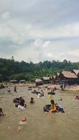 The video shows a group of people, mostly children, swimming and playing in a shallow, murky river. Several individuals are using inflatable tubes as flotation devices. The riverbanks are lined with rustic huts with thatched roofs. In the background, lush green trees cover a hillside. The sky is partly cloudy. Some people appear to be sitting in the huts or on the riverbank. The overall scene depicts a recreational activity in a natural environment.