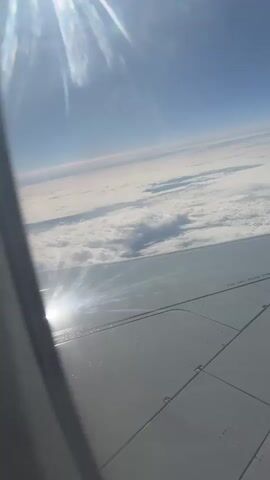 The video shows a view from an airplane window. The sky is a clear blue, and below the plane are clouds. The wing of the airplane is visible in the foreground. The sun is shining, creating a glare on the wing. The numbers "55868" are printed on the wing.