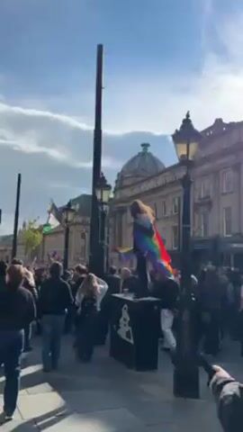 The video depicts a crowd of people gathered in a town square on a sunny day. A person stands atop a raised black platform, draped in a rainbow flag.  Several people in the crowd hold Palestinian flags. The crowd appears to be part of a demonstration or protest.  The buildings in the background have a classic architectural style, and lamp posts line the street. The sun is bright and slightly obscured by a few clouds.