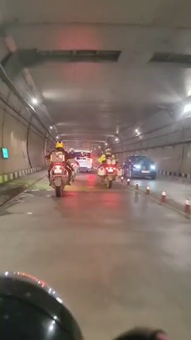 The video shows two motorcycles with riders, presumably in a tunnel, traveling in the same direction. The motorcycles are equipped with luggage cases on the rear. The riders are wearing helmets. There are other vehicles ahead, including a white car. The tunnel has overhead lighting and reflective markers along the sides. The road surface appears wet, and the red taillights of the motorcycles are reflected on the wet pavement. The camera is positioned behind the motorcycles, possibly from another motorcycle or a vehicle. The video is short, showing the motorcycles moving forward through the tunnel.
