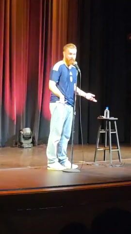 A man stands center stage at a microphone in a theater. He is wearing a dark blue t-shirt with white stripes and a circular emblem on the left chest, light blue jeans, and white sneakers. He gestures with his left hand while speaking into the microphone. To his right, a tall stool stands with a bottle of water and a small object on it. Behind him, red curtains are draped, and a stage light is visible on the floor to the left. The stage itself is made of polished wood.