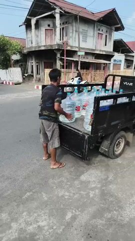 A man is unloading large plastic water jugs from the back of a small black truck. The truck is parked on a paved street in front of buildings. The man is wearing a patterned t-shirt and shorts. He is carrying two water jugs, one in each hand. The jugs are partially filled with water. In the background, there are other people and a motorcycle. The sun is shining, and the sky is clear.