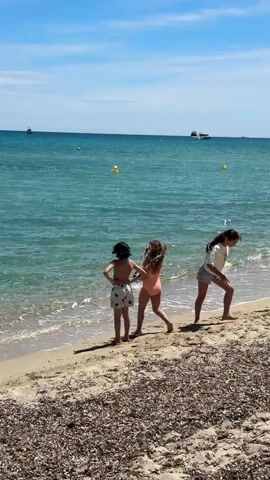 On a sunny day, three children are at the beach. Two of them, a girl in a salmon-colored swimsuit and a boy in patterned shorts, are standing at the water’s edge. The girl is holding what appears to be swim goggles. The third child, a girl in a light-colored top and shorts, is walking away along the beach. The water is a clear turquoise color and there are a few yellow buoys floating in the distance along with two small boats. The beach is sandy, with some seaweed or algae scattered on the sand closest to the camera.