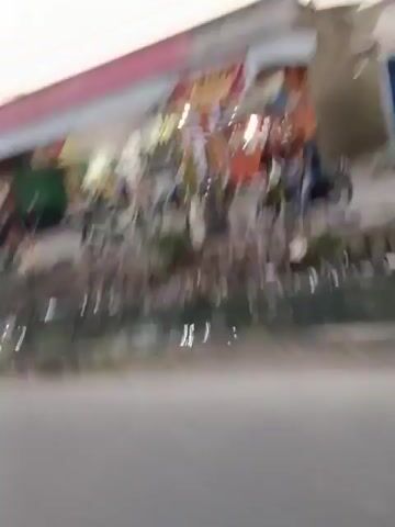 The video shows a street scene with several buildings lining both sides of the road. There are multiple motorcycles visible, some parked and others being ridden. People are seen interacting with the motorcycles and the environment. The buildings appear to be shops or residences, with some exhibiting signage. The road is paved, and there are utility poles and wires overhead. The lighting suggests it is either early morning or late afternoon. The overall impression is a moderately busy street in what appears to be a residential or commercial area.