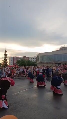 The video shows an outdoor performance by a group of dancers in traditional dark blue and red costumes, possibly performing a folk dance. They are moving in unison on a paved surface that appears wet, suggesting recent rain. A crowd of spectators is gathered behind the dancers, observing the performance. In the background, there are multi-story buildings and a tall evergreen tree, indicating an urban or town setting. The sky is overcast with a hint of sunset light breaking through the clouds. Some members of the audience are holding drinks. The camera angle is from a low perspective, focused on the dancers.