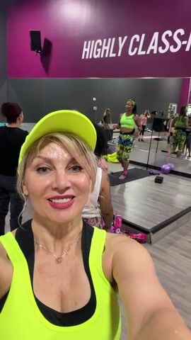 The video is a first-person selfie taken in a gym. A blonde woman in a neon yellow tank top and baseball cap is in the foreground, smiling at the camera. Behind her, a group of women are participating in a fitness class. They are in various poses, and some appear to be following an instructor who is clapping. The gym has mirrored walls and a large pink and white sign that reads "HIGHLY CLASS-IF." Yoga mats and other exercise equipment are visible in the background.