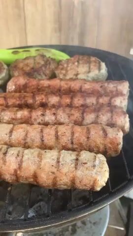 The video shows a grill filled with several items, including cooked cevapi, small hamburger patties, green peppers, and an onion. The cevapi are arranged in rows, showing grill marks. Smoke is rising from the grill. The background consists of a wooden fence.