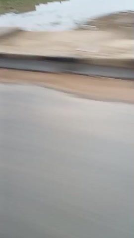The video shows a blurred perspective of a roadside guardrail and pavement, implying movement, possibly from a vehicle. The guardrail is white with black and silver segments and has a yellow line below it. Beyond the guardrail, a body of water or wet ground is visible. The pavement is gray and appears to be a road. The motion blur indicates a dynamic scene, potentially travel.