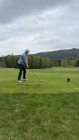A man wearing a light blue shirt, dark pants, white golf shoes, and a white baseball cap is playing golf on a green, grassy course. The sky is overcast. He swings a golf club and hits a golf ball off a tee. Another golf club is on the ground near his feet. In the background are trees and a mountain.