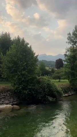 A body of water flows in the foreground, its surface rippling. On the left bank, a rocky embankment is visible, leading to lush green trees and bushes. Beyond the trees, a gentle slope covered in grass and scattered trees rises towards distant mountains. The sky above is partly cloudy with hints of sunset colors. The scene is serene and natural.