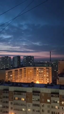 The video presents an elevated view of a cityscape at dusk. Several multi-story buildings are visible, with many windows illuminated, indicating activity within. The sky features a gradient of colors, ranging from a deep blue above to a pinkish hue on the horizon. Cables are visible in the foreground, stretching across the upper part of the frame. The area below the buildings is covered in snow.