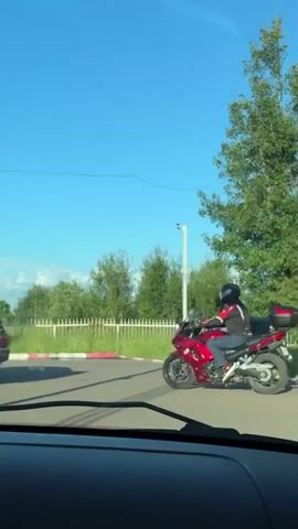 A person wearing a helmet and a dark jacket rides a red motorcycle with a black top box in a paved area under a clear blue sky. The motorcycle has a silver exhaust pipe and black wheels. The rider is wearing blue jeans and black shoes. The motorcycle is positioned on the right side of the frame, facing left. In the background, there is a white container or building, a tall white pole with a light fixture, trees, a fence with red and white markings, and a large black dumpster. A portion of a car can be seen on the left side of the frame. The lower part of the video shows the interior of a vehicle, likely the perspective of the person filming.