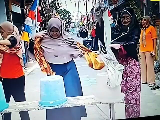 The video opens on a street scene with four women and several children. The women are dressed in hijabs and modest clothing. The first woman, on the left, wears a red shirt and black pants, holding a light-colored scarf. The second woman wears a light brown hijab, a blue skirt, and holds an orange scarf. The third woman wears a black hijab and patterned dress, holding a white scarf. The fourth woman, on the right, wears a black hijab, a patterned skirt, and holds a white scarf. Two blue buckets are positioned in front of the women. Children are visible sitting on the side of the road, watching. The scene appears to be part of a community event or celebration, with flags and decorations in the background. The women are engaged in an activity that involves the scarves and buckets, possibly a game. The camera then pans to show the women walking in a line across a crosswalk. They are all holding their phones and appear to be recording or taking pictures. More children are visible on the side of the road. The video then zooms in on the first three women again as they stand on the crosswalk, holding their phones. The woman on the left is wearing a yellow and brown scarf. The second woman wears a black and white patterned dress, and the third woman wears a white shirt and gray pants. Their attention is directed forward. The final shot shows the three women on the crosswalk, still holding their phones, with a bottle on the ground in front of them. The overall atmosphere seems festive and informal.