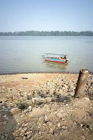 A small, multi-colored boat is seen from a rocky shoreline. The boat appears to have people on it. It is moving away from the shore and into a large body of water. The water is calm and reflects the clear sky. In the distance, a line of trees forms the horizon. The day is bright and sunny.