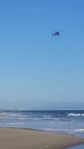 A police helicopter hovers in a clear blue sky above a beach. The helicopter is dark with white lettering that reads "LAPD AIR SUPPORT". Waves crash on the sandy shore below, where a few people are visible, including a person in white shorts and a dark shirt, and another person in dark shorts. The helicopter appears to be in motion, with its blades spinning. The scene suggests a day at the beach with a law enforcement presence overhead.