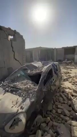 The video shows a car crushed under the rubble of a collapsed building. The car is gray and partially covered in debris. The building behind it is heavily damaged, with large cracks running through the remaining walls. Bricks and other construction materials are scattered around the car. The scene is bathed in bright sunlight under a clear blue sky. The overall atmosphere is one of destruction and devastation.