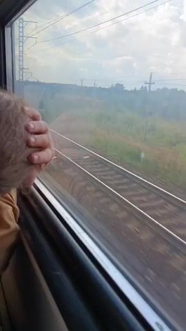 The video shows a view from a moving train window. The camera is positioned to capture the side of the train and the scenery outside. In the foreground, a hand, likely a person's, rests on the window frame. The window shows the reflection of the train interior on the left side. The view outside consists of train tracks stretching into the distance, bordered by grassy fields and vegetation. Overhead, power lines and utility poles are visible against a cloudy sky. The motion blur suggests the train is traveling at a moderate to high speed. The overall impression is a typical train journey through a rural or semi-rural landscape.