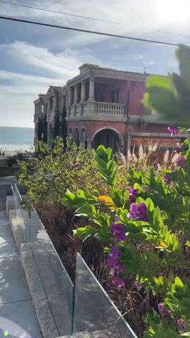 The video shows a view of a large, multi-story, red-colored building with classical architectural elements, including columns and balconies. The building appears to be situated near a beach and the ocean, which are visible in the background. In the foreground, there is lush green foliage with purple flowers, and a glass railing. The sky is partly cloudy. The camera appears to be positioned on a pathway or elevated area overlooking the scene.