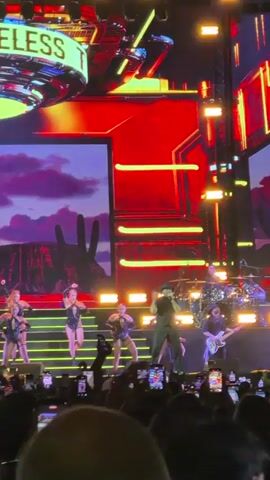 A male singer in a black hat and outfit performs on stage, center-right, with a group of female dancers in matching black outfits to his left. A band, including a drummer and guitarist, is visible behind them. The stage features large screens displaying a desert landscape with saguaro cacti under a purple and pink sky. Above the screen, a large circular illuminated sign reads "ICELESS TIMBER". The concert is well-lit with red and yellow stage lights, and the audience in the foreground is holding up numerous smartphones to record the performance.