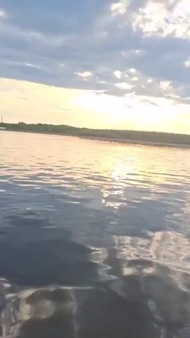 A first-person perspective shot of a body of water at sunset. The sun is low on the horizon, casting a warm glow across the sky and reflecting on the water's surface. Clouds are visible in the sky, with some sunlight breaking through them. The horizon line is marked by a strip of trees. The water is slightly rippled, indicating a gentle movement.