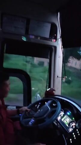 The video begins by showing the interior of a vehicle, likely a truck or bus, from the perspective of the driver. The dashboard, steering wheel, and windshield are visible. Outside the windshield, a rural landscape is visible, with houses, trees, and power lines. The video then transitions to an external view of the vehicle as it drives along a paved road. The road appears to be in a rural area with vegetation and some scattered houses. The sky is overcast, suggesting it may be dusk or a cloudy day. The vehicle's headlights are on, illuminating the road ahead. The video ends with the vehicle continuing down the road.