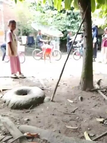 The video shows an outdoor scene with several children and adults. In the foreground, a child wearing a pink shirt and patterned pants is visible, standing next to a large, circular, stone object in the dirt. The child appears to be looking downwards. In the background, other children are seen riding bicycles. One child in a pink shirt and pants is standing and looking towards the camera. Another child in a red shirt is on a bicycle. An adult in a black shirt is also on a bicycle. Further in the background, there are buildings and trees. The ground is covered in dirt, with some scattered sticks and leaves. The overall atmosphere appears to be casual and outdoors, possibly a residential area.