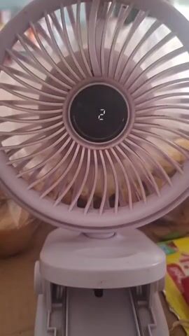 A purple clip-on fan is shown in a close-up shot. The fan has a circular guard with thin spokes radiating from the center. In the center of the guard is a small black display showing the number 2 in white digital font. The fan is mounted on a white base with a clip mechanism. The background is blurry but appears to be a tabletop with some objects on it, including what looks like bags and possibly some packaging. The video is composed of static frames, indicating no motion.