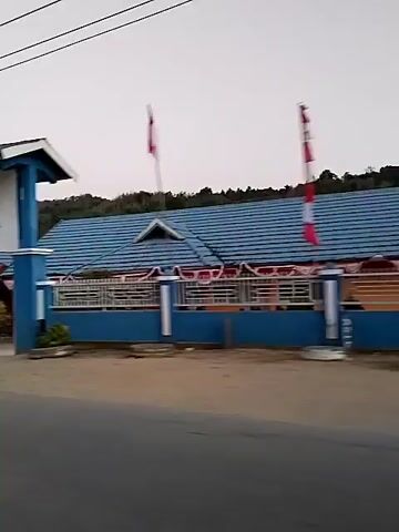 The video pans across the exterior of a school building. The first frame shows the entrance gate with the school's name visible: "SMP NEGERI 20 POLEANG UTARA KABUPATEN BOMBANA". The building has blue roofs and orange and white walls. Flags are visible. In the second frame, people in headscarves are seen near the fence and flags. The video continues to pan, showing more of the school grounds and surrounding area, including trees, houses, and a hilly landscape in the background. The sky is overcast.