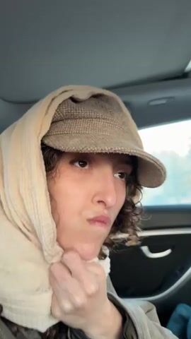 A woman is filmed from a front-right perspective, seated in the passenger seat of a car. She has curly brown hair and is wearing a beige baseball cap underneath a light-colored, possibly cream or off-white, headscarf that covers her head and part of her neck. She is also wearing a dark, possibly olive-green or brown, jacket. Her right hand is visible, holding the headscarf near her chin.

The woman exhibits a range of expressions, including talking, smiling, and a slight pout. The car appears to be in motion, as the background visible through the side window is blurred. The lighting suggests it is daytime, possibly early morning or late afternoon, with soft, ambient light entering the vehicle. The interior of the car, including parts of the headliner and the door, is visible. At the very beginning of the video, faint Hebrew text briefly appears on screen, reading "סופר הכוונה" (Super Intention). The video's location context is Mitzpe Ramon, Israel.