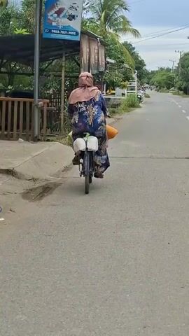 A person wearing a blue floral print dress and a beige hijab rides a bicycle down a paved road. The person is carrying two white containers and an orange bag on the back of the bicycle. The road is lined with buildings, vegetation, and a sidewalk. The sky is cloudy. The video appears to have been filmed outdoors during the daytime.