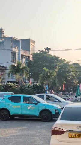 The video shows a street in Vietnam with cars, buildings, and palm trees. In the foreground, there are several cars, including a blue "Xanh SM" taxi with black rims, a white car, and other vehicles. The taxi has a phone number, 1900 2068, displayed on its side. Behind the cars, there are buildings of varying heights, with the tallest one appearing to have multiple levels and a satellite dish on top. Palm trees and other greenery are interspersed between the buildings. The sky is a hazy pale blue, suggesting either early morning or late afternoon lighting. Power lines run across the top of the frame. There is some activity in the background with people and vehicles, including what appears to be a row of market stalls or vendors along the side of the road. Overall, the scene appears to depict a normal, busy urban street.