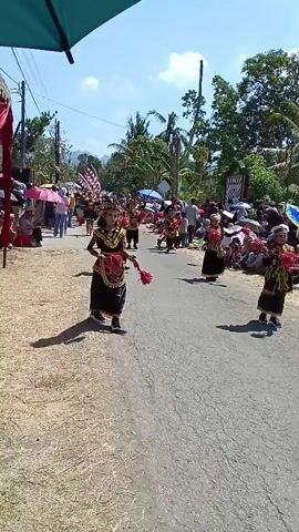 A young girl in traditional Indonesian attire, including a headdress and a black and gold costume, performs a dance on a paved road lined with spectators. She holds two red fans, one in each hand, and moves with her arms extended. The girl is walking forward while dancing. The sun is shining brightly, casting shadows on the road. Other children in similar costumes are visible further down the road, also participating in the procession. The spectators, some holding umbrellas, are seated and standing on the sides of the road. Trees and buildings can be seen in the background. The scene appears to be a cultural event or festival.