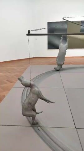 This video captures a dynamic art installation within a museum setting. The primary subjects are two sculptures of animals, appearing to be dogs, rendered in a matte gray material. One dog is depicted in a low, crouched posture, with one foreleg extended and a trail of dark pigment suggesting movement on the floor. The second dog is suspended upside down from a wire system attached to a central, tripod-like structure. This structure supports a mechanical element, likely a motor, and a series of taut wires that extend outwards, creating a tension and kinetic possibility within the artwork.

The installation is situated on a large, flat, gray floor marked with grid lines and circular traces. In the background, a large, abstract painting with muted tones of black, gray, and olive green hangs on a white wall. Further in the distance, another piece of art, a wooden shelving unit with three cubbies, is mounted on the wall. The overall setting is a well-lit, spacious museum gallery with polished wooden flooring visible around the edges of the main exhibition area.

The video pans across the installation, showcasing the interplay between the suspended and ground-based sculptures, the mechanical elements, and the surrounding artworks. The lighting suggests it is daytime, and the indoor environment implies controlled climate conditions. The artwork evokes a sense of motion, perhaps a chase or a dance, and the precariousness of the suspended figure adds an element of drama. Two individuals are briefly visible in the background, observing the art, but their presence is secondary to the main installation. No visible text is present in the video. The location is identified as The Hague, Netherlands.