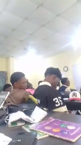 The video shows a classroom environment with several students seated at desks. The camera is positioned at a low angle, likely from one of the students' desks. In the foreground, a table is visible with a purple book, a phone, and a calculator. The students are mostly facing away from the camera, engaged in what appears to be a class or study session. One student in the center is wearing a black and white shirt with the number "35" on the back. Another student to the left is wearing a brown and white checkered shirt. In the background, a whiteboard is partially visible, with some writing on it. The room has a bright ceiling light and light-colored walls, suggesting a typical classroom setting.