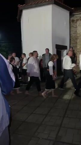 The video captures a night scene in Balgari, Bulgaria, where a group of people are walking outdoors on a paved surface next to a building. Many individuals are barefoot, particularly those dressed in what appears to be traditional attire, including white shirts and dark skirts or trousers. Other people are observing or walking alongside in casual clothing. The movement suggests a procession or a ritualistic walk, possibly related to the local Nestinarstvo tradition, which involves barefoot dancing. Traditional Bulgarian folk music, featuring a gaida (bagpipe) and percussion, plays in the background, setting a cultural atmosphere. The building has white plastered walls on one section and a stone facade on another, with a red-tiled roof. Artificial lighting illuminates the scene, indicating it is nighttime.