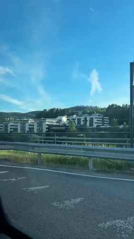 The video shows a view from a moving vehicle, likely a car, looking out the passenger side window. The scene is an outdoor environment on a clear day with a bright blue sky and scattered white clouds. In the background, there are several modern buildings with white facades, some with multiple stories and balconies, nestled on a gently sloping green hill covered with trees. A metal guardrail runs along the side of the road in the foreground. Beyond the guardrail, there is a strip of dry grass and then a body of water, possibly a river or lake, with more trees lining the opposite bank. The vehicle appears to be on a road or bridge elevated above the water and the surrounding landscape. The sunlight creates reflections on the windows of the buildings and casts shadows on the road. A tall pole, possibly a lamppost or utility pole, is visible on the left side of the frame. The video seems to capture a scenic drive through a suburban or semi-rural area with architectural elements.