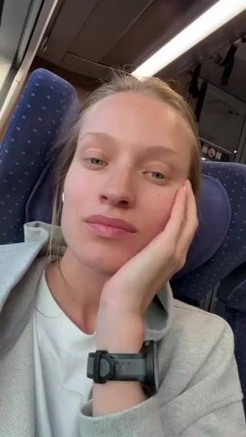 A woman with blonde hair and blue eyes, wearing a grey hooded jacket, a white shirt, and a black digital watch on her left wrist, is seated on a train. She has an earbud in her right ear and is adjusting it with her right hand. The train seats are blue with white headrests. The sun shines on her face through the window. She looks to her right and then directly at the camera.