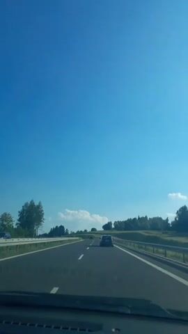 The video shows a first-person view from inside a car driving on a highway. The sky is a clear, vibrant blue, suggesting a sunny day with good weather. The road ahead is paved and appears to be a multi-lane highway, indicated by the white lines marking the lanes and the guardrails on the sides. 

The car is driving on the right side of the road. In front of the car, another dark-colored car is traveling in the same direction. Further ahead, there are hints of other vehicles, suggesting moderate highway traffic. 

The surrounding landscape is characterized by rolling hills covered in lush green trees and foliage. There are also some scattered white clouds in the distance, adding to the scenic view. The time of day appears to be afternoon, given the bright sunlight and the angle of the shadows. 

The overall mood is peaceful and relaxing, typical of a scenic drive. There are no people visible outside the cars, and the focus is on the journey and the open road. The license plate of the car in front is partially visible, but the text is not clear enough to be read. The location is identified as Rdzawka, Poland.