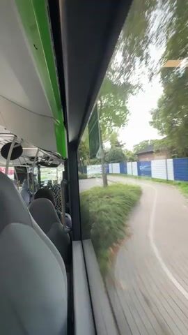 This video shows the view from inside a moving bus, looking out the window on the right side. The camera is angled slightly forward. In the foreground, we see the backs of several gray bus seats. The window is large, and through it, we see a blurred green landscape with trees. A sidewalk or path runs parallel to the bus, and beyond that, there is a blue fence and a white sign. The sky is overcast. The bus appears to be traveling through a suburban or park-like area.