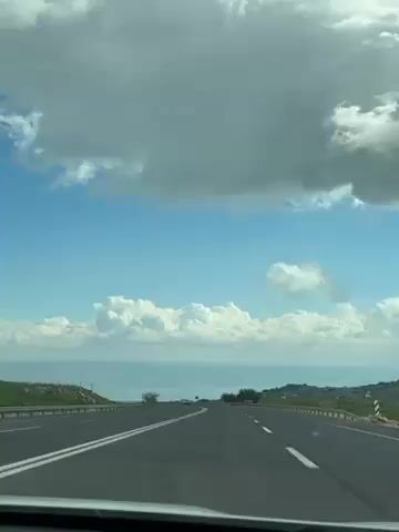 The video shows a view from inside a car driving on a highway towards a body of water under a partly cloudy sky. The road is paved with white lane markers. Other vehicles are visible in the distance. The sky is predominantly blue, with patches of white clouds. On either side of the road, there is vegetation. The water appears to be a lake or sea, with hills or mountains visible in the background. A parking sign is visible on the right side of the road.