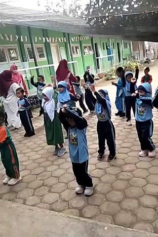 The video shows a group of young children and a few adults standing outside on a paved area. The children appear to be engaged in some sort of synchronized activity, possibly a dance or exercise routine, as they are all facing the same direction and making similar movements with their arms and bodies. Many of the children are wearing blue uniforms with headscarves, while others are in different colored outfits. The adults, also dressed in headscarves and modest clothing, seem to be supervising the children. In the background, there is a building with green siding and windows, suggesting an outdoor school or community setting. The ground is covered with light-colored hexagonal paving stones. The overall atmosphere appears to be one of a structured educational or recreational activity for young children.