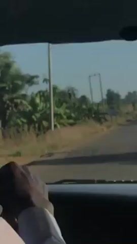 A car is shown driving down a narrow, paved road, past a roadside signpost. The view is from inside the car, capturing the steering wheel and the driver’s hand. The signpost indicates "MANCHOCK 11 KM" to the right and "MARARRUN" and "KAGORO 10KM" to the left. The environment appears rural with vegetation along the roadside. Overhead powerlines are visible. The sky is clear.