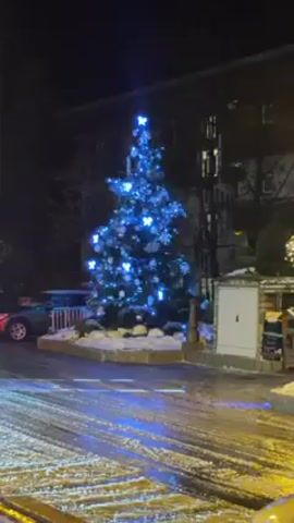 The video depicts a Christmas tree illuminated with blue lights at night. The tree is decorated with snowflake ornaments and has a star on top. It stands on a small patch of ground covered in snow, next to a street covered in snow and slush. Cars can be seen parked nearby, and a street lamp casts a warm glow on the scene. The surrounding buildings are visible in the background, but they are dark and difficult to discern in detail.